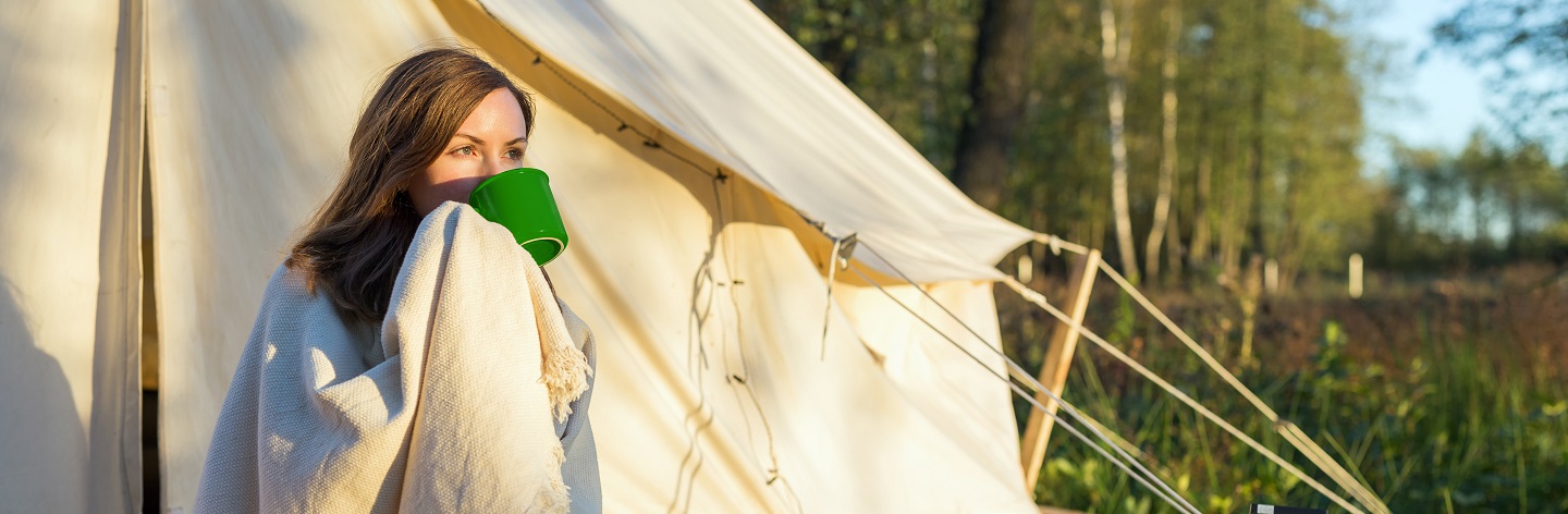 Young Woman Wraps Blanket Over Herself While Drinking Coffee Near Canvas Tent In The Morning