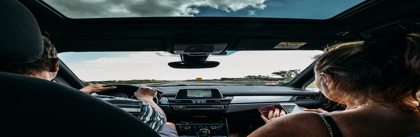 Older Couple On Front Seat Of Car. Road Trip Concept. Woman Uses A Smartphone To Pass The Time While Man Drives